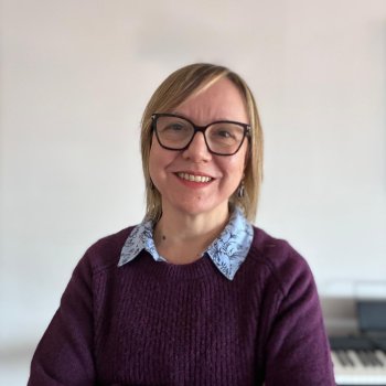 Emel Memiş’in açık renk tenli, kısa sarı saçlı ve siyah çerçeveli gözlüklü portre fotoğrafı. Gülümsüyor ve kolları göğsünde çapraz duruyor. Üzerinde koyu mor bir kazak var; yakasından açık mavi, desenli bir gömlek yakası görünüyor. Arka plan çoğunlukla düz, açık renk bir duvar; sağ alt tarafta bulanık şekilde bir klavye/piyano benzeri enstrüman.