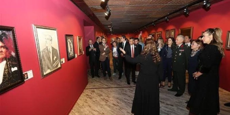 Kırmızı duvarlarla çevrili bir sanat galerisi veya müze koridorunda bir grup insan. Duvarlarda çeşitli portre resimleri asılı. Kalabalık, bir kadının rehberliğinde resimleri inceliyor. Kadın, siyah bir elbise giymiş ve gruba bir şeyler anlatıyor. Grubun içinde resmi kıyafetli kişiler de var, bunlar arasında bir askeri üniforma giymiş bir adam dikkat çekiyor. Zemin ahşap görünümlü ve tavanda spot ışıkları yer alıyor.