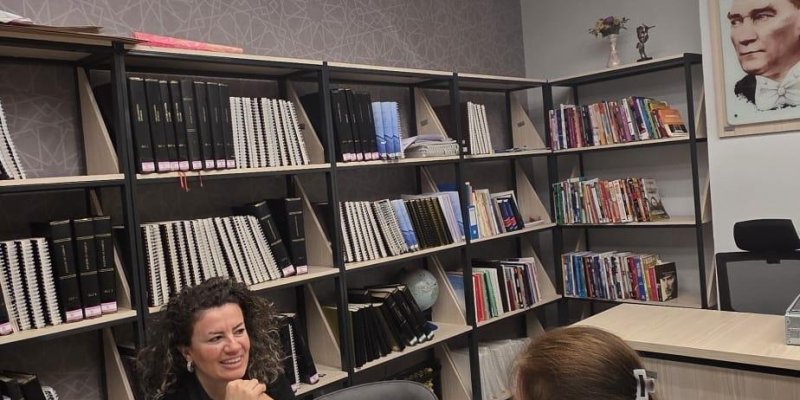 Bir kütüphane ya da çalışma odasında, masanın iki yanında oturan Emine Kamçı ve Hilal Yıldırım. Biri kameraya yakın tarafta sırtı dönük şekilde oturuyor; uzun kahverengi saçlı ve saçında toka var, yeşil desenli bir üst giymiş. Karşısındaki koyu kıvırcık saçlı, siyah giyinmiş; dirseklerini masaya koyup ellerini çenesine dayamış ve gülümsüyor.