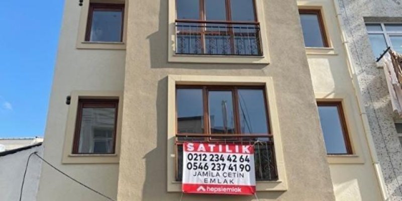 Bej ve krem tonlarında dört katlı bir apartmanın dış cephesi. Ortada iki balkonda siyah ferforje korkuluklar ve kahverengi çerçeveli cam kapı/pencereler görünüyor; yanlarda daha küçük pencereler var. Orta kattaki balkonda kırmızı-beyaz “SATILIK” yazılı bir afiş asılı; altında telefon numaraları yazıyor, “EMLAK” ifadesi seçiliyor. En üstteki balkon korkuluğunda da benzer bir afişin üst kısmı kısmen görünüyor. Gökyüzü açık mavi, bina cephesine güneş vuruyor.