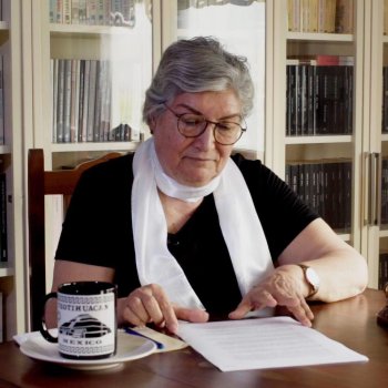 Kitaplıkların önünde bir masada oturuyor. Kısa, gri saçlı ve gözlüklü; siyah bir üst ve boynunda beyaz bir atkı/şal var. Başını hafifçe öne eğmiş, masadaki basılı bir kâğıda odaklanmış; bir eliyle kâğıdı tutuyor, diğer eliyle satırları takip ediyor. Masanın sol tarafında tabak içinde bir kupanın üzerinde “MEXICO” yazısı. Masanın sağ tarafında altın renkli, fil başlı bir heykel (Ganeşa figürü) duruyor. Heykelin yanında bir gözlük ve masanın sağ alt köşesinde mavi kenarlı bir kitap/defter. Arka pl