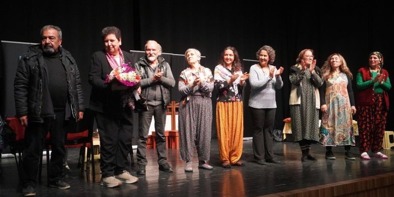 Sahnede bir grup insan yan yana duruyor. Sol başta siyah mont giymiş bir adam, yanında, elinde çiçek buketi tutan ve siyah takım elbise giymiş bir kişi, ortada, deri ceket giymiş yaşlı bir adam, onun yanında, desenli elbise giymiş birkaç kadın sıralanmış. Kadınlardan bazıları ellerini çırpıyor. En sağda, kırmızı yelek ve yeşil bluz giymiş bir kadın var. Sahne arka planı siyah ve sahnede birkaç sandalye bulunuyor.
