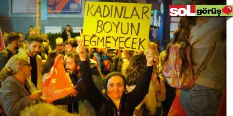 Kalabalık bir grup insanın katıldığı bir protesto veya gösteri. Ön planda, yaşlı bir kadın sarı bir pankart tutuyor. Pankartta büyük harflerle "KADINLAR BOYUN EĞMEYECEK" yazıyor. Kadının yüzünde kararlı bir ifade var. Etrafında Türk bayrağı taşıyan başka insanlar da bulunuyor. Arka planda çeşitli binalar ve tabelalar görülüyor. Fotoğrafın sağ üst köşesinde "sol görüş" yazısı.