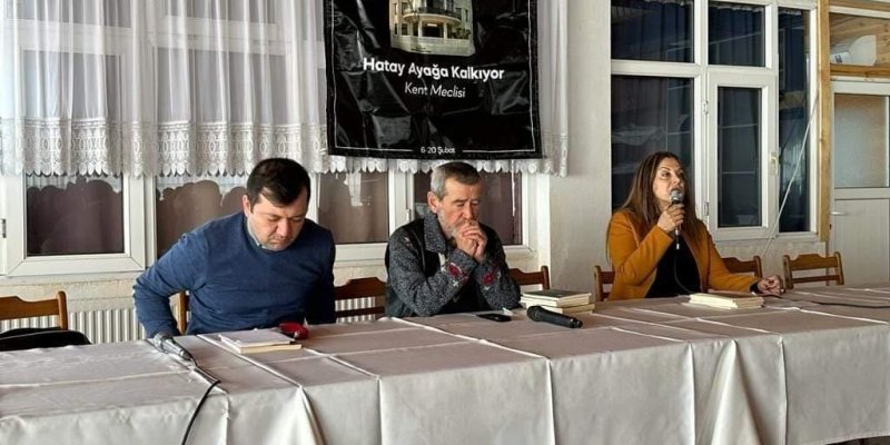 Bir masa etrafında oturan üç kişi. Masanın üzerinde beyaz örtüler ve mikrofonlar yer alıyor. Soldaki adam mavi bir kazak giymiş ve önünde bir mikrofon var. Ortadaki adam gri bir kazak giymiş ve ellerini çenesine dayamış. Sağdaki kadın turuncu bir ceket giymiş ve elinde bir mikrofon tutuyor, konuşuyor gibi görünüyor. Arkada, duvarda "Hatay Ayağa Kalkıyor Kent Meclisi" yazılı bir afiş asılı. Mekân, beyaz perdeler ve ahşap tavan ile dekore edilmiş.