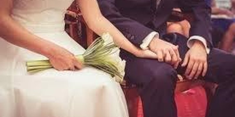 Kırmızı sandalyede oturan, başları ile dizlerinin üzerleri görüntülenen gelin ve damat var. Damat lacivert takım giyinmiş. Gelinin bir eli damadın dizi üzerinde damadın elini tutuyor. Gelinin diğer eli dizinin üzerinde ve bir buket beyaz çiçek tutuyor.