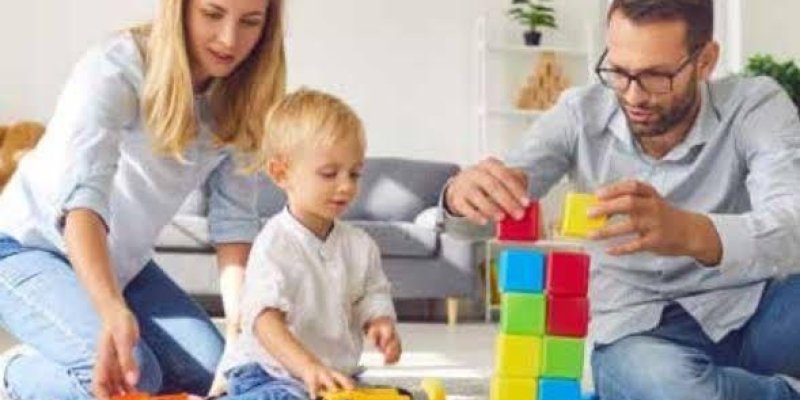 Bir aile oturma odasında yerde oturmuş ve birlikte oyun oynuyor. Solda, sarı saçlı bir kadın, mavi kot pantolon ve açık renk bir üst giymiş. Ortada, sarı saçlı küçük bir çocuk, beyaz bir tişört giymiş. Sağda, gözlüklü bir adam, mavi kot pantolon ve açık renk bir gömlek giymiş, renkli bloklardan bir kule yapıyor. Çocuk ve kadın oyuncak kamyonlarla oynuyor. Arka planda, gri bir kanepe ve bitkilerle süslenmiş bir raf görülüyor.