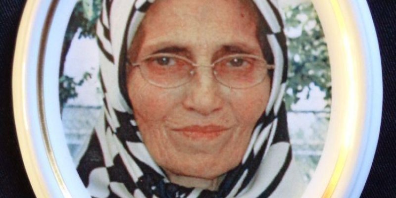 Fikriye Kodak, siyah ve beyaz desenli bir başörtüsü takmış, gözlüklü, orta yaşlı, yüzünün yakın çekimi görülüyor. Hafif gülümseyerek kameraya bakıyor.