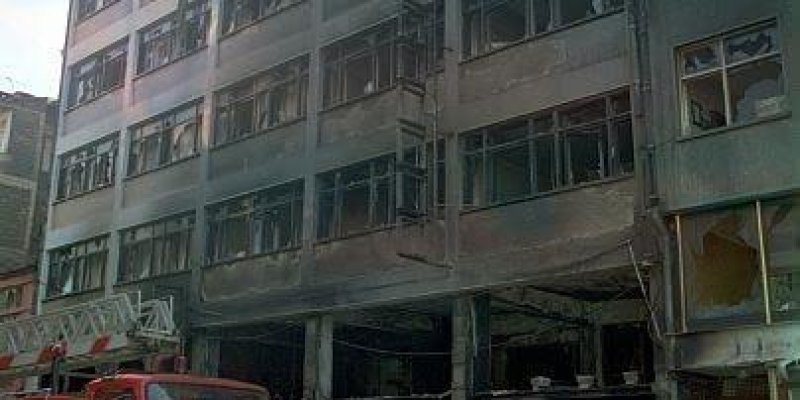 Yanmış bir apartman cephesi. Beş katlı, gri renkli büyük bir bina; cephenin tamamında yoğun yanık izleri, is ve kararmış yüzeyler. Özellikle pencerelerin çevresi koyu kararmış, bazı camlar kırılmış veya tamamen yok, bazı çerçeveler dışarı doğru eğilmiş. Alt kattaki dükkân veya giriş kısmı ciddi hasar almış; kolonlar isli ve kararmış, içeri doğru boş ve karanlık, kapı boşlukları açık. Binanın önünde yerde yanmış enkaz yığınları, kömürleşmiş malzemeler ve dağınık metal parçaları. Sol tarafta kırmı