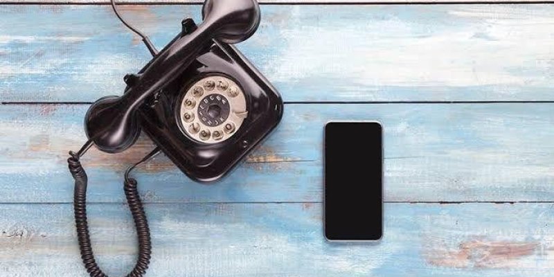 Eski tip bir siyah döner numaralı telefon ve modern bir akıllı telefon yan yana duruyor. Zemin, mavi ve beyaz tonlarında ahşap bir yüzeyden oluşuyor. Eski telefonun kablosu kıvrımlı ve ahize kısmı telefonun üzerinde duruyor. Akıllı telefonun ekranı kapalı ve siyah renkte. İki telefon, eski ve yeni teknolojiyi temsil eder şekilde yan yana yerleştirilmiş.