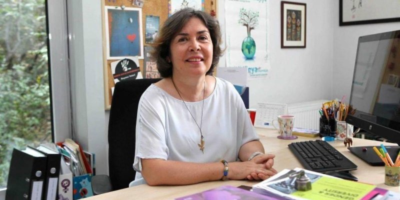 Bir ofis masasının arkasında oturan, gülümseyen Şemsa Özar’ın teni açık, saçları kısa ve koyu kahverengi; beyaz kısa kollu bir bluz giymiş ve boynunda kolye var. Masanın üzerinde klavye, bilgisayar ekranı, kalemliklerde kalemler, bir kupa bardak, çeşitli dosya ve evrak yığınları duruyor. Sol tarafta pencere yanında klasörler ve dosyalar var. Arkadaki panoda kartpostallar ve notlar asılı; “foruma ekolojije” yazılı bir poster ve duvarda çerçeveli yazı/tablolar bulunuyor.