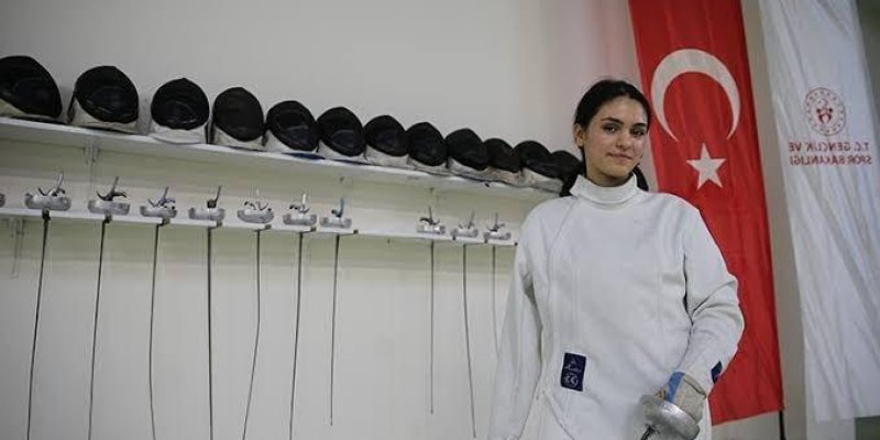 Meryem Hanne Erbil, eskrim kıyafeti giymiş genç bir sporcu. Beyaz eskrim tulumu ve eldiveni içinde, sağ elinde bir eskrim kılıcı (flöre ya da epe) tutuyor, uç kısmı yere doğru. Kısa koyu renk saçları geriye toplanmış, açık tenli ve hafifçe gülümsüyor. Arka planda dua sabit uzun bir raf üzerinde yan yana dizilmiş çok sayıda siyah eskrim maskesi ve maskelerin altına asılı kılıçlar, kılıçların ince uzun namluları aşağı doğru sarkıyor. Sağ tarafta büyük bir Türk bayrağı dikey şekilde asılı; yanında