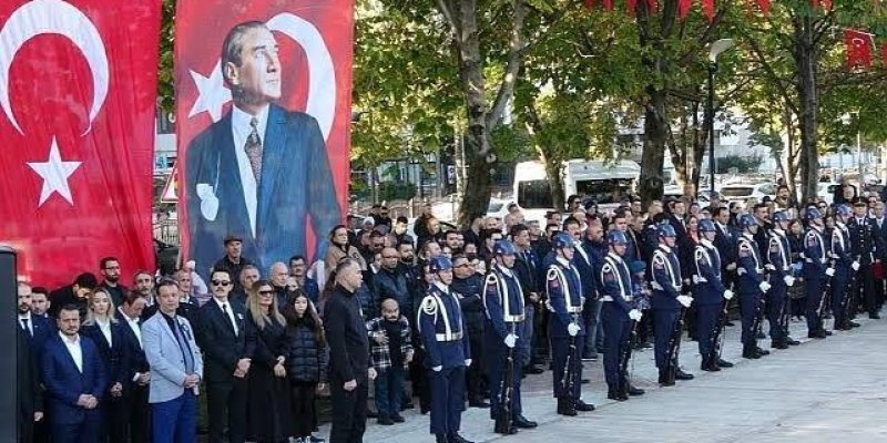 Açık havada düzenlenen bir tören. Sol tarafta, büyük bir Atatürk posteri ve Türk bayrakları asılı. Posterde Atatürk, takım elbise giymiş. Sağ tarafta resmi üniformalar giymiş bir grup asker sıralanmış. Askerlerin önünde ve arkasında sivil kıyafetli insanlar. Arka planda ağaçlar ve park etmiş araçlar. Tören alanı, geniş bir kaldırım üzerinde. Üst kısımda Türk bayrakları asılı.