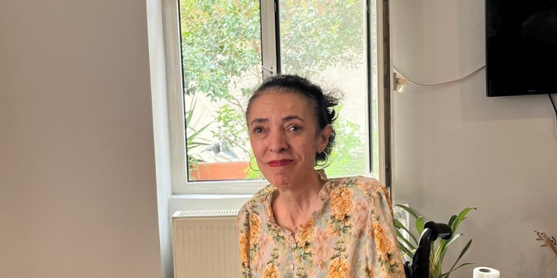 Fatma Çoban, masa başında oturuyor tekerlekli sandalyede. Saçları koyu renk ve geriye toplanmış. Üzerinde açık renk zeminli, turuncu çiçek desenli bir bluz; sol bileğinde açık renk boncuklu bileklik. Yüz ifadesi hafif gülümser gibi ve başı kameraya dönük. Ön planda ahşap bir Masanın solunda gri bir dizüstü bilgisayarın köşesi ve sarı bir bilgisayar faresi var; fareye bağlı siyah kablo masaya yayılmış. Masanın ortasına yakın bir yerde, üzerinde yazı olan bir kâğıt duruyor. Arka planda beyaz çerçe