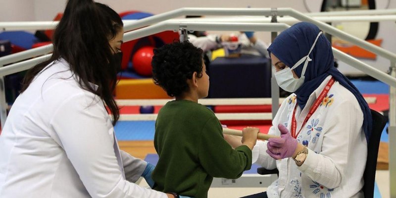 Kapalı bir rehabilitasyon/terapi alanında, iki sağlık çalışanının bir çocuğa egzersiz yaptırdığı bir sahne. Ortada yeşil kazaklı, kıvırcık saçlı küçük bir çocuk ayakta duruyor ve iki eliyle yatay bir tutamağı kavrıyor. Sağ tarafta oturan, lacivert başörtülü ve cerrahi maskeli bir kadın mor eldiven takmış; çocuğun ellerini yönlendiriyor ve boynunda kırmızı bir yaka kartı askısı var. Sol tarafta beyaz önlüklü, siyah maskeli bir kadın mavi eldiven takmış; çocuğun bel/sırt kısmından destek veriyor.