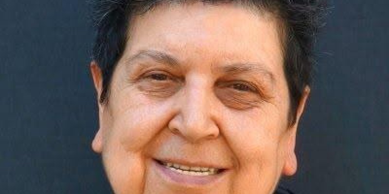 Şenal Saruhan’ın kısa, koyu renkli, kabarık saçlı portresi. Ten rengi buğday ile açık esmer arasında. Kaşları koyu renkli ve seyrek, gözleri hafif kısık. Sol gözünün altı biraz daha koyu gölgeli. Dudakları kapalı, hafif gülümseyen, sakin ve sıcak bir ifade. Üzerinde koyu renkli bir giysi; yakası açık, iç kısmında daha koyu bir katman seçiliyor. Arka plan düz, koyu gri-siyah renkte.