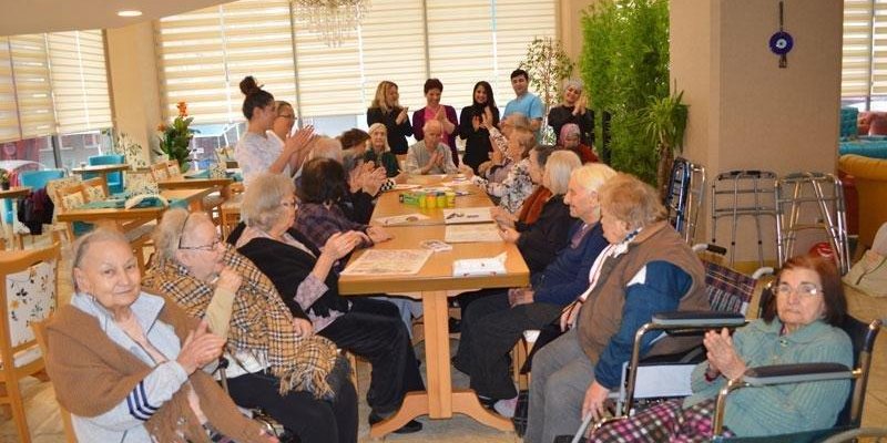 Yaşlıların bir arada bulunduğu, uzun masalı bir toplu etkinlik fotoğrafı. İç mekânda, aydınlık bir salon içinde iki yanda sandalyelerde ve tekerlekli sandalyelerde oturan çok sayıda yaşlı kişi; çoğunun saçları beyaz veya gri. Bir kısmı ellerini çırpıyor. Ortada dikdörtgen bir masa; masanın üzerinde kâğıtlar ve renkli küçük kutular duruyor. Masanın arka tarafında ayakta duran birkaç yetişkin kişi var; bazıları alkışlıyor. Arkada geniş pencereler boyunca açık renk stor perdeler görünüyor. Sağ tara