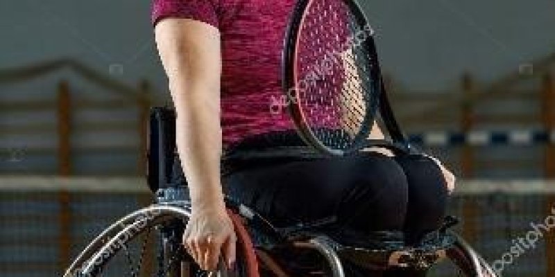 Tekerlekli sandalyede oturmuş gülümseyen genç bir kadın var. Kadının bir eli tekerleği tutuyor, diğer elinde de tenis raketi var.