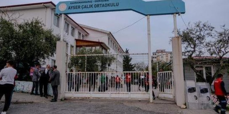 Demir parmaklıklı beyaz bir okul kapısı ve kapının üstünde mavi bir tabela. Tabelada büyük harflerle “KAHRAMANMARAŞ – DULKADİROĞLU AYSER ÇALIK ORTAOKULU” yazıyor. Kapının hemen içinde ve çevresinde birkaç yetişkin ve öğrenci grubu ayakta duruyor ve sohbet ediyor. Arkada beyaz renkli, birkaç katlı, kırmızı çatılı bir okul binası uzanıyor; uzun dikdörtgen pencereler sıralı. Kapının yanlarında ve arka tarafta ağaçlar. Zeminde gri, hafif yıpranmış bir asfalt alan.
