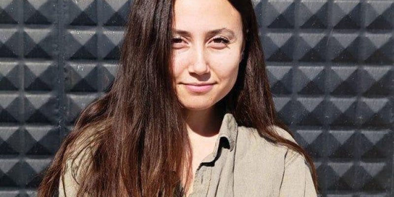 Esra Işık, uzun kahverengi saçlı, kare kabartmalı koyu gri bir duvarın önünde duruyor. Yüzü doğrudan güneşe dönük, aydınlık; gözleri kısık ve dudaklarında hafif, kapalı bir gülümseme. Cildi açık tenli, yanaklarında güneşten hafif bir kızarıklık. Saçları ortadan ayrılmış, düz ve omuzlarının altına kadar uzanıyor, sağ tarafı öne doğru düşmüş. Üzerinde haki renginde, uzun kollu, göğsünde cepleri olan bir gömlek bulunuyor; en üst düğmesi açık. Arka plandaki duvar, düzenli kare çıkıntılardan oluşan d