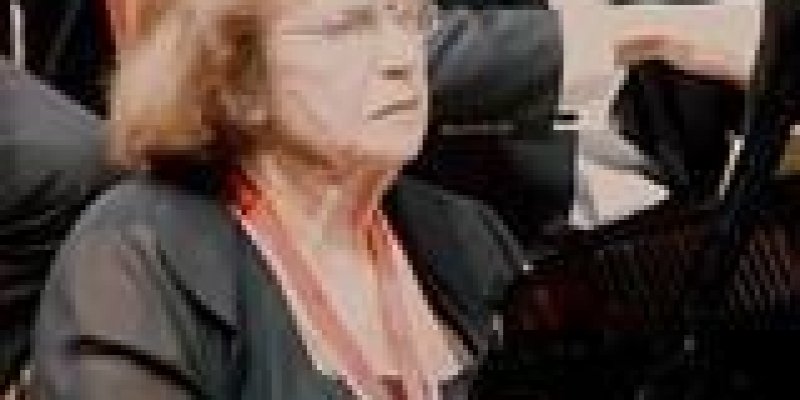 Piyanist Ayşe Gül Sarıca’nın 60-65 yaşlarında fotoğrafı. Sahnede, piyanonun başında oturur şekilde, arkasında orkestra, keman çalan bir kişi. Sarıca siyah şifon kıyafetli, beyaz inci kolyeli, kumral, küt saçlı, gözlüklü.