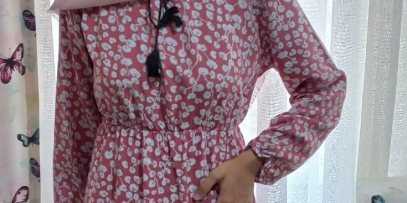 Pembe ve beyaz renkli çiçek desenli bel kısmında siyah bir kemer olan elbise giymiş, başında pembe bir eşarp olan bir kadın, beyaz tül perde önünde duruyor ve hafif sağa dönük bir şekilde gülümsüyor. Arka planda sol tarafta kelebek desenli bir perde ve sağ tarafta bir pencere görünüyor.