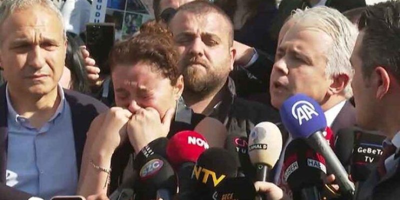 Kalabalık bir basın açıklaması sahnesi. Ön planda sağda gri saçlı bir erkek, mikrofonlara doğru konuşuyor. Ortada bir kadın ağlıyor; gözleri kapalı, elleriyle yüzünü kapatmış. Solda başka bir erkek ciddi bir ifadeyle kameraya bakıyor. Kadının ve konuşan erkeğin önünde çok sayıda mikrofon var; üzerlerinde AA ve NTV gibi logolar seçiliyor. Arkada kalabalık ve bazı kişilerin elinde telefonlar/kameralar görülüyor.