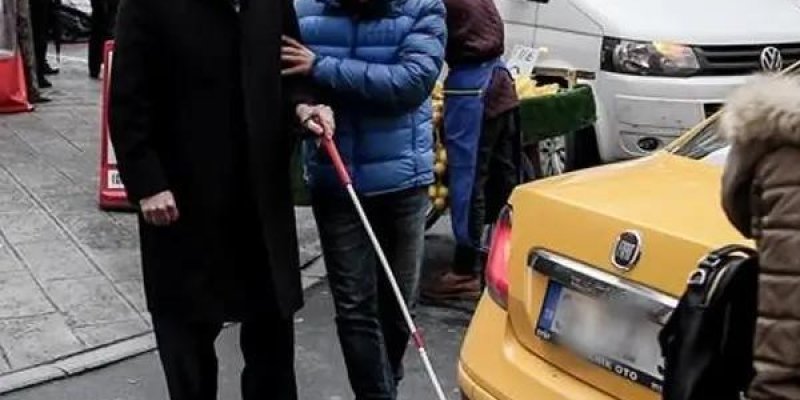 Kaldırım kenarında duran iki erkek ve ön planda sarı bir taksi. Yaşlı erkek gri saçlı, açık tenli, uzun siyah palto ve koyu renk kıyafetler giymiş; yüzünde gülümseme ifadesi var. Yanındaki daha genç erkek açık tenli, kısa koyu saçlı ve sakallı; mavi şişme mont, kot pantolon ve açık renk bot giymiş. Genç erkeğin elinde kırmızı saplı beyaz bir baston var; baston ucu yerde ve öne doğru uzatılmış. Sarı taksinin arka kısmı kadrajın sağında; arka stop lambası ve bagaj kapağı görünüyor, plaka kısmı bul