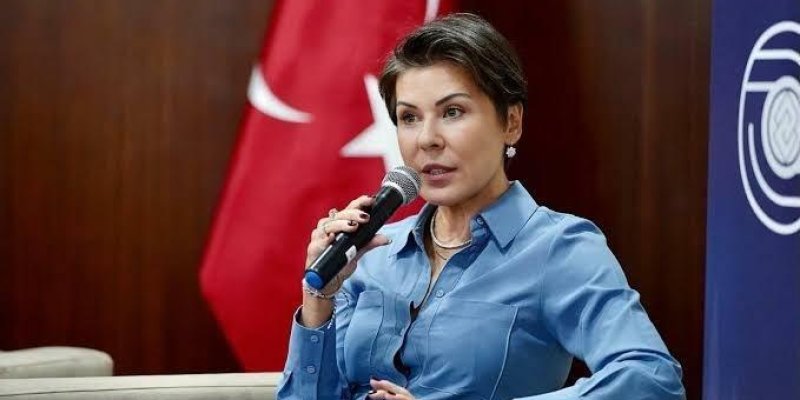 Pınar Türker, kısa koyu kahverengi saçlı, açık tenli, elinde mikrofonla konuşuyor. Mavi, uzun kollu, düğmeli bir gömlek giymiş, bileğinde saat veya akıllı bileklik, boynunda zincir kolye, kulaklarında küçük küpeler, bir kolunu koltuğun kolçağına yaslamış, diğer eliyle mikrofonu yüzüne yakın tutuyor; ifadesi ciddi ve odaklanmış. Oturduğu yer açık renk, modern bir koltuk ve sahne ya da panel ortamı. Arka planda büyük bir Türk bayrağının kırmızı zemininde beyaz ay-yıldızın bir kısmı. Sağ tarafta ma