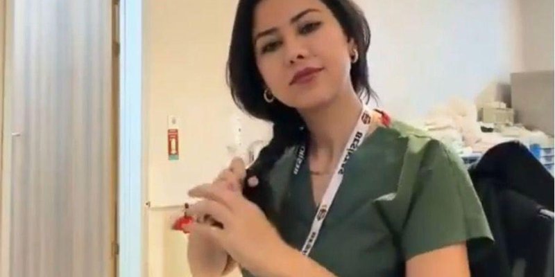 Yeşil forma giyen bir kadın, iç mekânda kameraya bakarken saçını elleriyle yan tarafa doğru topluyor. Boynunda yazılı bir yaka kartı askısı var ve kulağında halka küpeler görünüyor. Arka planda ofis ya da klinik benzeri alanda bir sandalye ve bazı eşyalar seçiliyor.