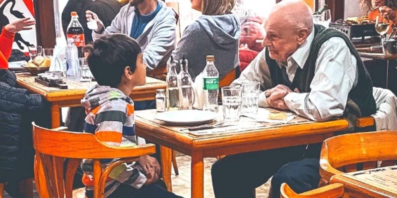 Ahşap sandalyeli küçük bir restoran masasında karşılıklı oturan yaşlı bir adam ve küçük bir çocuk. Çocuk, renkli yatay çizgili (turuncu, gri, beyaz ve yeşil tonlarında) kapüşonlu bir sweatshirt giymiş, masaya dönük şekilde yaşlı adama bakıyor. Yaşlı adamın başında saç çok az, teni açık renk, beyaz gömlek ve koyu yeşil süveter yelek giymiş, dirseklerini masaya dayamış, ellerini birbirine yakın tutarak çocuğa doğru eğilmiş. Masada boş beyaz tabaklar, birkaç cam bardak, cam şişelerde yağ, soslar ve