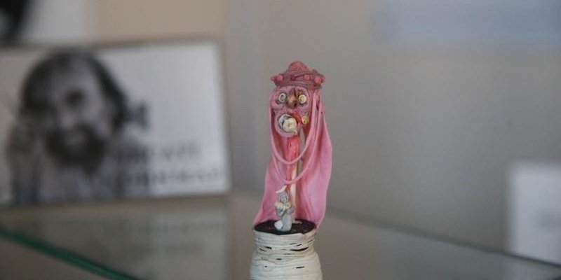 Cam bir raf üzerinde duran küçük bir heykelcik, pembe bir pelerin giymiş ve yüzü maskeli bir figürü tasvir ediyor. Figürün yüzü oldukça detaylı ve maskesi dikkat çekici. Heykelciğin alt kısmı siyah ve beyaz renklerde, sarmal bir desenle süslenmiş. Arka planda, bulanık bir şekilde, bir kişinin siyah-beyaz fotoğrafı.