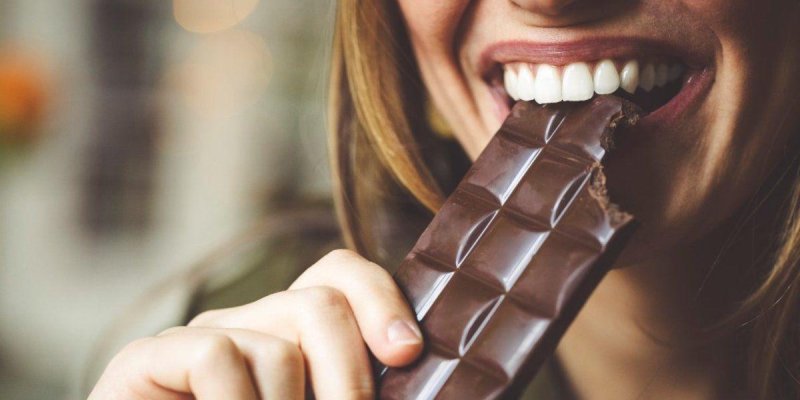 Bir kişinin ağzının ve elinin yakın plan fotoğrafı; kişi bir parça bitter çikolatayı ısırıyor. Çikolatanın üstünde kare dilimler belirgin ve bir köşesinde ısırık izi var. Kişinin dişleri görünüyor ve dudakları hafif gülümser gibi. Arka plan bulanık ve iç mekân ışıkları gibi yumuşak, yuvarlak ışık lekeleri seçiliyor.