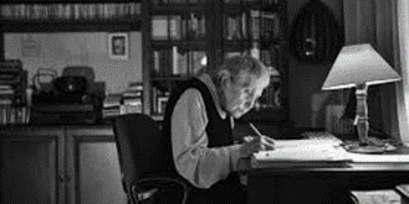 Siyah beyaz bir fotoğrafta orta yaşlı bir kadın, büyük ve klasik bir çalışma masasının başında oturuyor. Kadının önünde açık bir kitap ve kağıtlar var ve o, ciddi bir ifadeyle çalışmaya odaklanmış, bir şeyler yazıyor. Masanın üzerinde bir masa lambası var ve ışık doğrudan kağıtların üzerine düşüyor. Arka planda kitap dolu bir kitaplık ve bir telefon görülüyor. Oda klasik bir dekorasyona sahip.