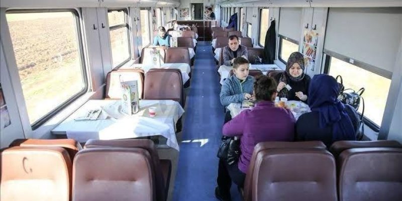 Tren vagonunda yemekli bir bölüm. Masalarda beyaz örtüler. Bazı masalarda insanlar oturuyor. Ön planda, üç kadın ve bir çocuk masada yemek yiyor. Kadınlardan biri mor bir ceket, diğerleri koyu renkli giysiler giymiş. Arka planda başka masalarda da oturan insanlar var. Vagonun pencerelerinden dışarısı görünüyor, tren hareket halinde.