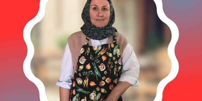 Rahime Söylemez Çetin, başörtülü ve beyaz bir gömlek ve üzerinde çeşitli sebze desenleri olan siyah bir önlük giymiş. Fotoğrafın kenarları dalgalı beyaz bir çerçeve ile çevrelenmiş ve arka plan kırmızı tonlarında.