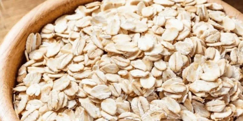 Tahta bir kâse içinde açık bej renkli yulaf ezmesi. Kâse ağzına kadar dolu ve yulaf taneleri yassı, pul pul görünüyor. Kâse ahşap bir yüzey üzerinde duruyor. Arka tarafta solda buğday başakları, sağ üstte gri-beyaz bir kabın kenarı kısmen görünüyor.