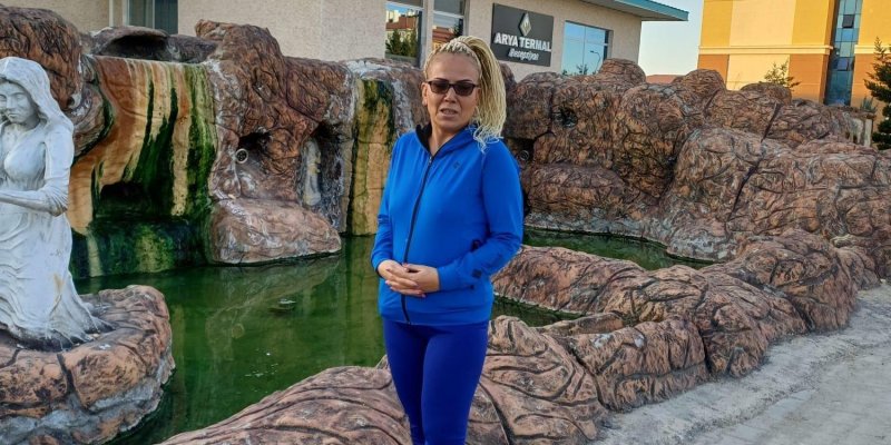 Döndü Kanat, açık havada mavi bir eşofman üstü ve tayt giymiş, siyah spor ayakkabılı. Gözlerinde güneş gözlüğü var ve sarı saçları omuzlarına kadar uzanıyor. Arka planda, yapay bir kaya formasyonu, küçük bir su havuzu. Kaya formasyonunun yanında beyaz bir heykel var.