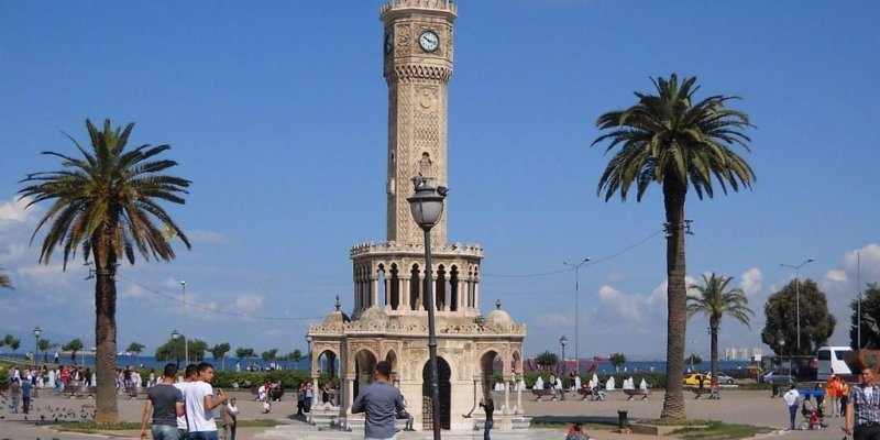 İzmir’in simgelerinden biri olan Kordon’daki Saat Kulesi. Yazın güneşli bir günde Kule’nin arkasında deniz, iki yanında birer tane palmiye ağacı ve meydanda birçok insan görünüyor.