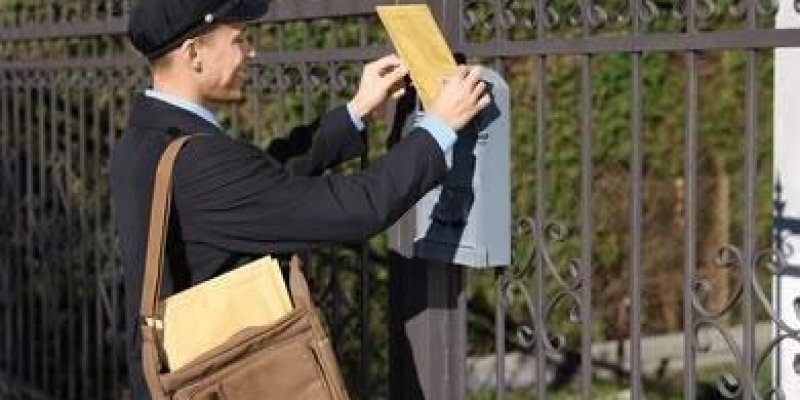 Klasik postacı kıyafeti giymiş bir adam, demir parmaklıklı bir çitin üzerindeki posta kutusuna zarf atarken. Adam koyu renk bir ceket, açık mavi gömlek ve koyu renk bir şapka takmış. Omzunda kahverengi deri görünümlü bir postacı çantası asılı ve çantanın içinde büyük sarı zarflar. Elinde tuttuğu sarı zarfı, gri renkli dikdörtgen bir posta kutusunun kapağından içeri yerleştiriyor.