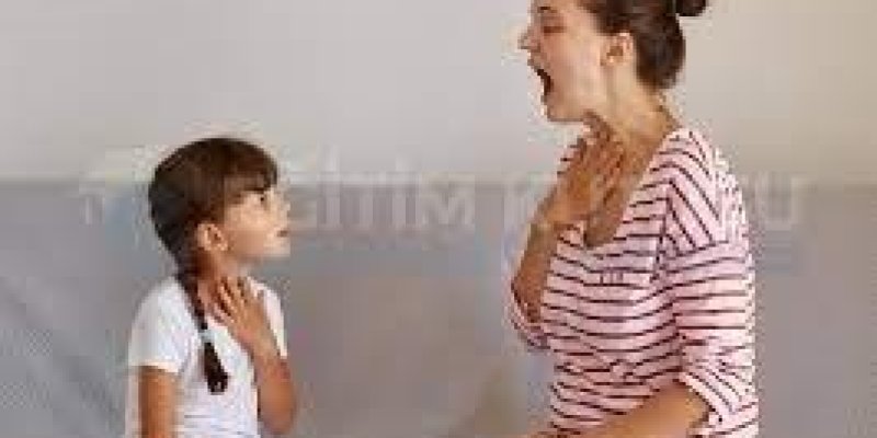 Kanepede yan yana oturmuş ve bedenleri birbirine dönük 5-6 yaşlarında bir kız çocuğu ve yetişkin bir kadın var. Kadın tek elini kendi boğazına götürmüş ve ağzını açmış bir şekilde kız çocuğuna bakıyor. Çocuk da kendi elini boğazına götürmüş ve kadını izliyor. İşitme engelli kız çocuğuna dudak okuma eğitimi veriliyor.