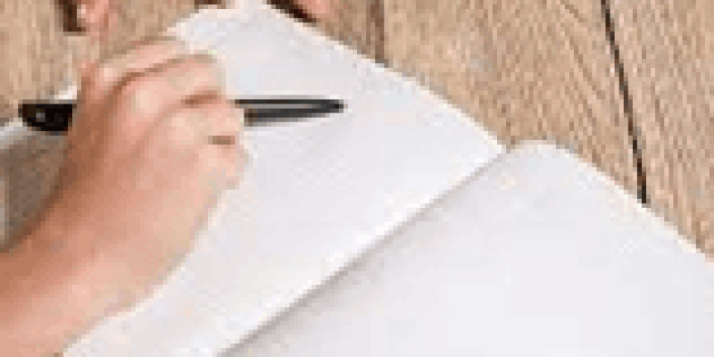 Ahşap bir masanın üzerinde açık bir defter var. Defterin sağ sayfasında bir kişi kalemle yazı yazıyor. Arka planda, defterin sol üst köşesinde bir kitap, kitabın sadece arka kapağı ve bir kısmı görünüyor, kapakta herhangi bir yazı veya resim yok. Kişinin sol eli defterin sol sayfasının altında, sağ eli ise yazı yazarken kalem tutuyor.