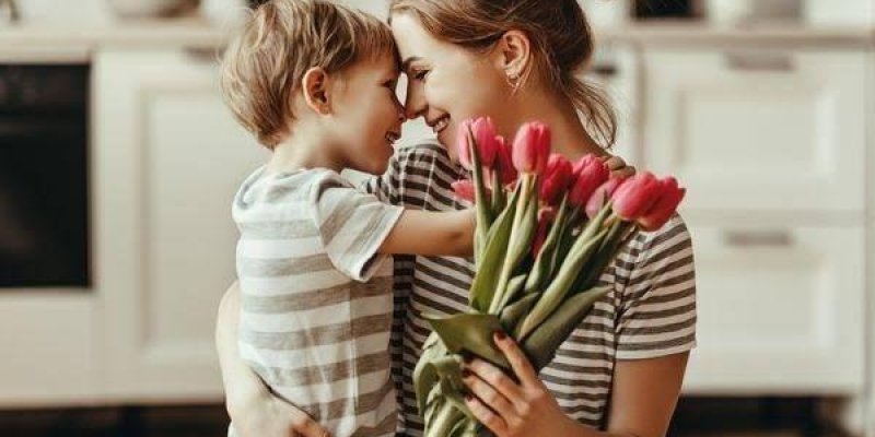 Bir anne ve küçük oğlu mutfakta birbirlerine sarılmış. İkisi de çizgili tişört giymiş. Anne, elinde bir demet kırmızı lale tutuyor. Anne ve oğul birbirlerine çok yakın duruyor ve burun buruna gelmişler. Arka planda bulanık bir mutfak tezgâhı ve dolaplar var.