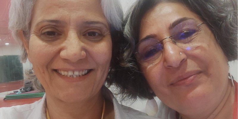 Sultan Çamur Karataş ve Nursel Denir beyaz veya gri saçlı, gülümseyerek kameraya bakıyorlar. Yapay Zekam: İki kadın yakın çekim bir selfie çekmiş. İkisi de beyaz veya gri saçlı ve gülümsüyorlar. Soldaki beyaz bir gömlek giymiş ve boynunda sarı bir yaka kartı var. Sağdaki gözlüklü, beyaz bir üst giymiş ve boynunda kırmızı bir yaka kartı taşıyor. Arka planda beyaz bir duvar ve sol üstte pembe bir alan görünüyor.