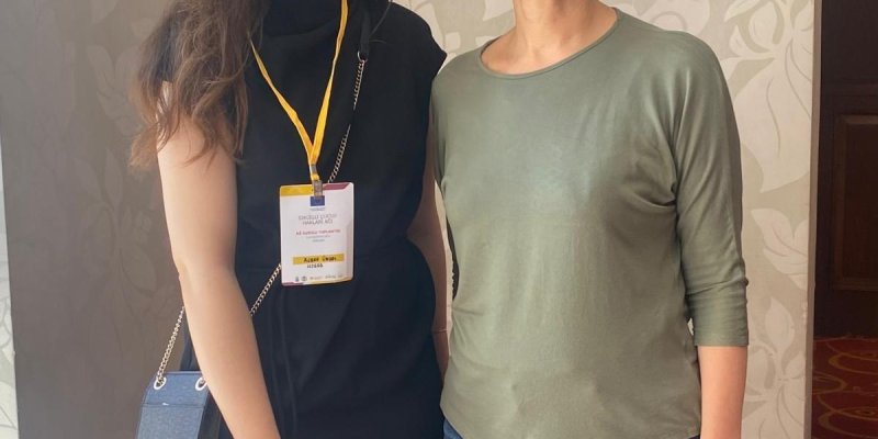Söyleşideki iki kadın yan yana duruyor. Soldaki uzun, dalgalı kahverengi saçlı ve siyah bir elbise giymiş. Boynunda sarı bir yaka kartı asılı ve elinde mavi bir çanta taşıyor. Sağdaki kısa, gri saçlı ve yeşil bir bluz ile mavi kot pantolon giymiş. İkisi de gülümsüyor ve arka planda açık renkli, çiçek desenli bir duvar kâğıdı var. Zemin kırmızı halı kaplı.