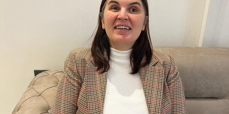 Renkli pötikareli ceketinin içindeki beyaz kazağı, omuz hizasındaki ortadan ayrılmış düz saçlarıyla bej rengi bir koltukta bir kadın gülümseyerek oturuyor.