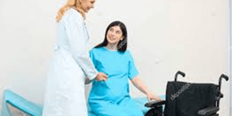 Bir doktor ve hamile bir kadın. Doktor beyaz bir önlük giymiş ve sarı saçlı. Hamile kadın mavi bir hastane elbisesi giymiş ve tekerlekli sandalyede oturuyor. Doktor, kadının yanında duruyor ve ona gülümsüyor. Arka planda bir muayene masası var. Fotoğrafın sol alt köşesinde "depositphotos" yazısı bulunuyor.