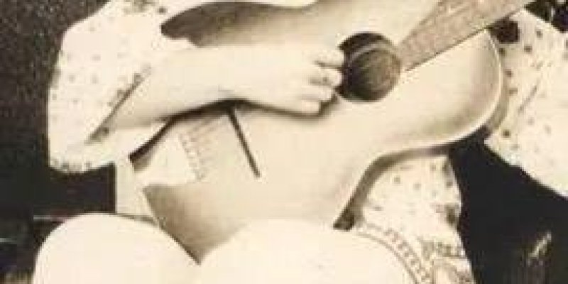 Ülkü Aker gitar çalıyor. Orta uzunlukta, tepede toplanmış kabarık bir saç modeli var; saçları açık renk. Açık desenli, rahat bir bluz ve açık renk geniş paçalı bir pantolon giymiş. Akustik gitara kucağında, gövdesi yana dönük şekilde sarılmış; sol eli sapın üzerinde, sağ eli gövdenin üzerinde duruyor. Yerde oturur pozisyonda, bacakları hafif çaprazlanmış. Arkada camlı bir kapı ve bitki yaprakları. Yüz ifadesi hafif ciddi ve düşünceli.