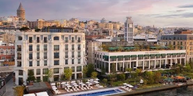 Deniz kenarında büyük bir otel kompleksi. Ön planda denize paralel uzanan uzun, dar bir yüzme havuzu ve etrafında beyaz şezlongların, güneş şemsiyelerinin dizildiği bir teras alanı. Havuzun hemen arkasında ağaçlar ve bitkilerle çevrili bahçe düzenlemesi. Solda 5 katlı, açık renk taş cepheli, dikdörtgen pencereli klasik görünümlü bir bina. Sağında, daha yatay uzanan, üst katında yeşil çatı bahçesi olan modern bir bina. Cephesinde sütunlar ve geniş camlar.