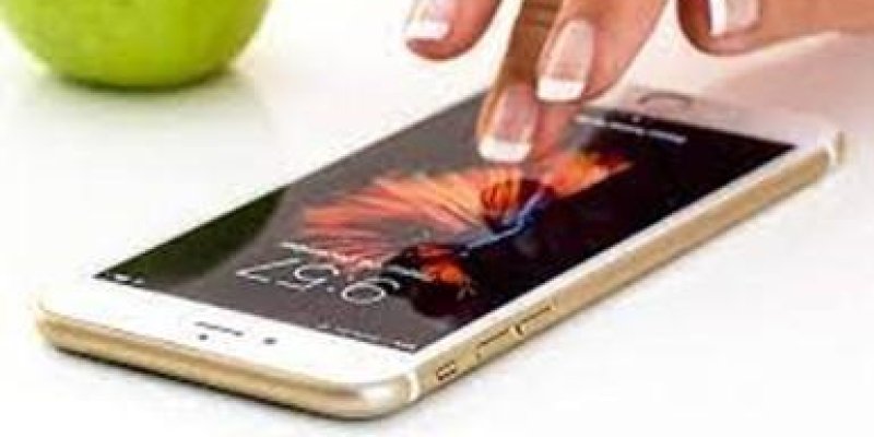 Beyaz zeminin üzerinde saat 9.57 yi gösteren akıllı telefon var. Telefonun fonunda alev rengi temalı duvar kâğıdı görünüyor. Telefonun üzerinde tırnaklarının ucu beyaz ojeli ve renksiz cilalı genç bir kadın eli var. En arka kısımda yeşil bir elmanın alt kısmı görünüyor.
