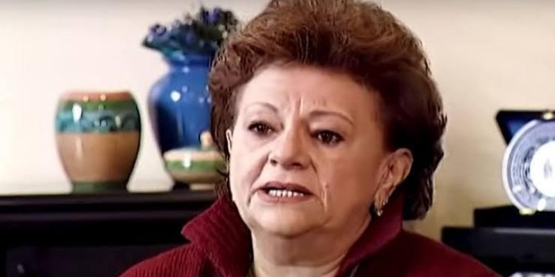 Kısa kahverengi saçlı ve kırmızı bir kazak giymiş yaşlı bir kadın. Yüz ifadesi ciddi ve dudakları hafifçe aralanmış. Arka planda, bir raf üzerinde duran mavi ve yeşil tonlarında iki vazo ve bir tabak bulunuyor. Kadın, iç mekân gibi görünen bir ortamda oturuyor.